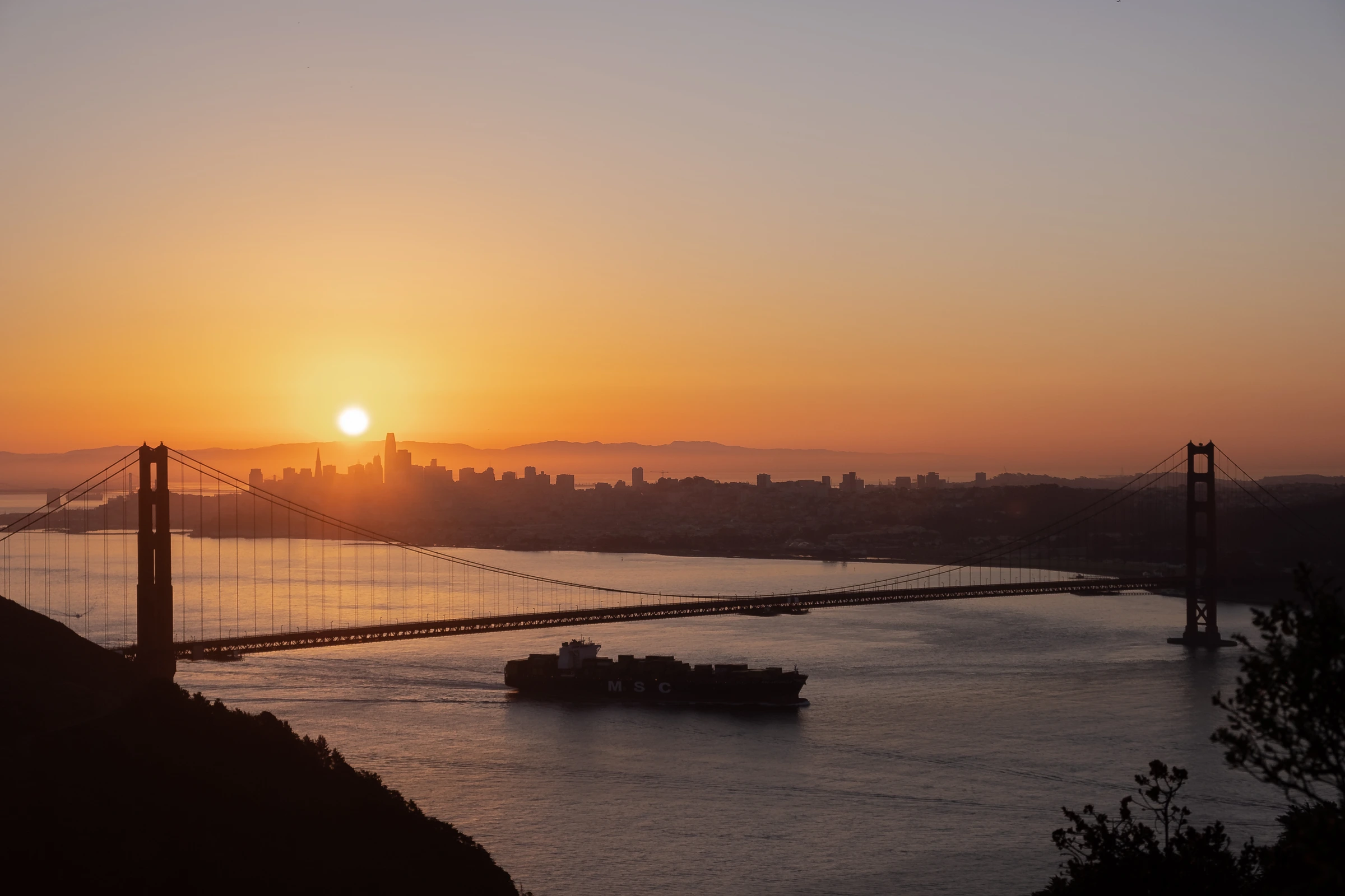 Golden Gate Bridge at sunrise, San Francisco skyline silhouetted, container ship passing beneath.