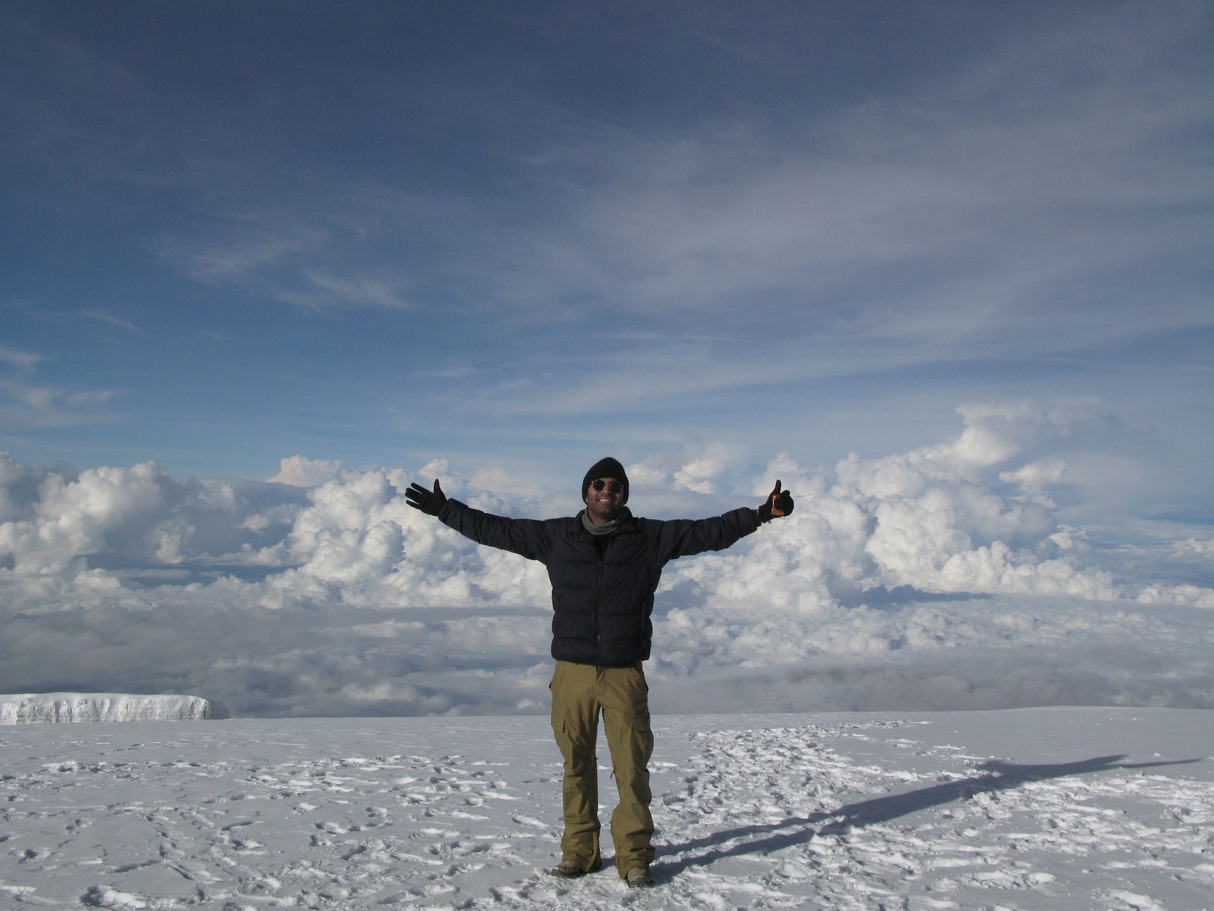 Amar at the Kilimanjaro summit, arms outstretched, snow underfoot, clouds below.