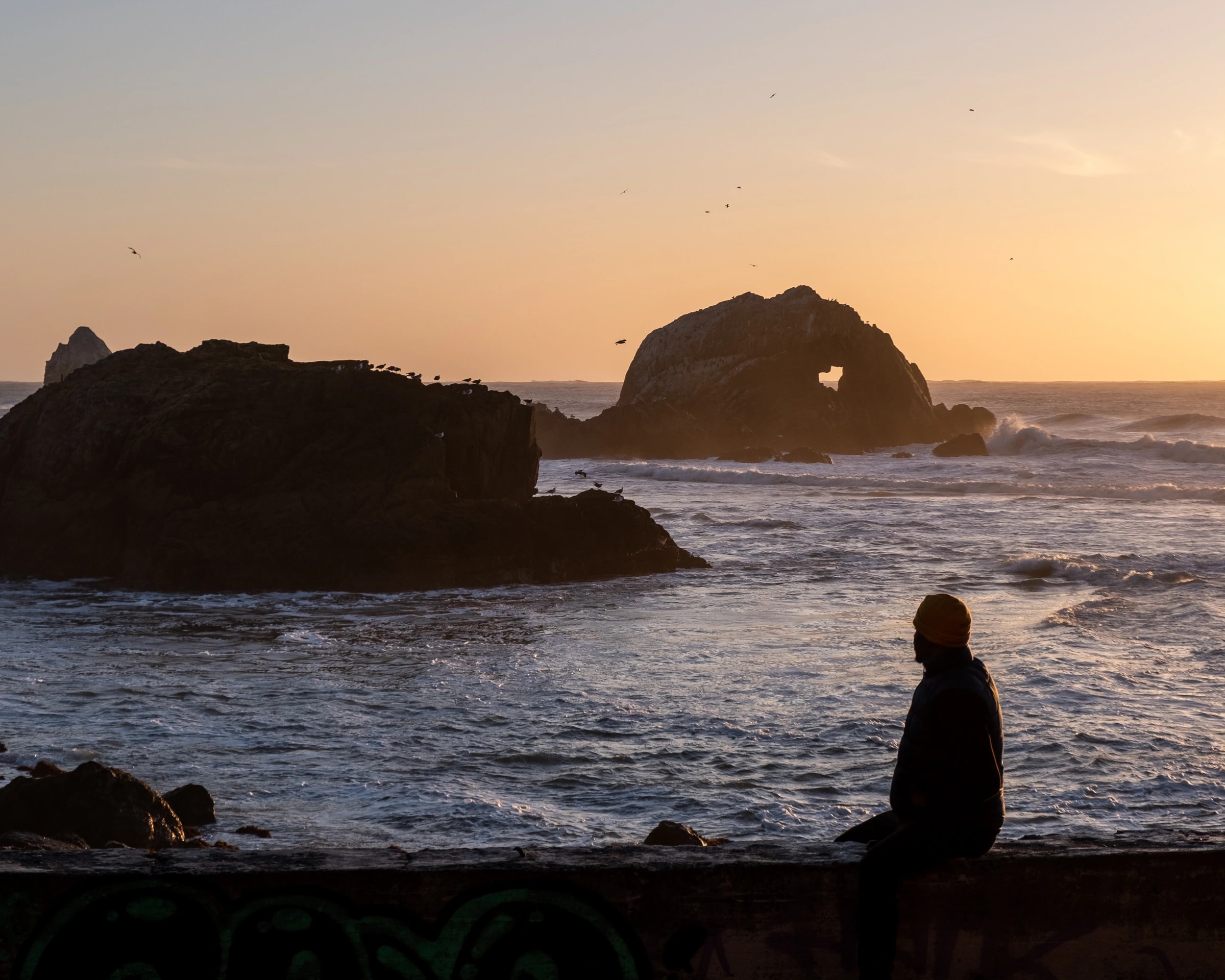 Person in a beanie on a graffiti-covered wall watching the Pacific at sunset, sea arch in distance.